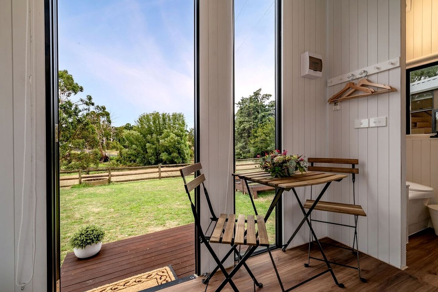Een kamer met een houten tafel en stoelen, een kleine plant in een pot en uitzicht op een grasveld door grote ramen.