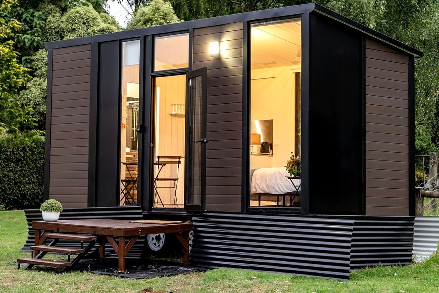 A compact, modern cabin with a sliding glass door entrance, a small patio with a table and chairs, and a visible interior with a bed and a lamp.