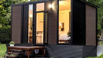A compact, modern cabin with a sliding glass door entrance, a small patio with a table and chairs, and a visible interior with a bed and a lamp.
