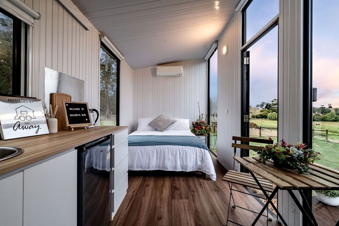 A bedroom with a bed, a small table, a bench, and a view of the outdoors.