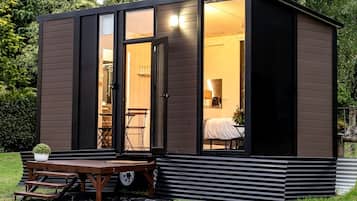 A compact, modern cabin with a sliding glass door entrance, a small patio with a table and chairs, and a visible interior with a bed and a lamp.