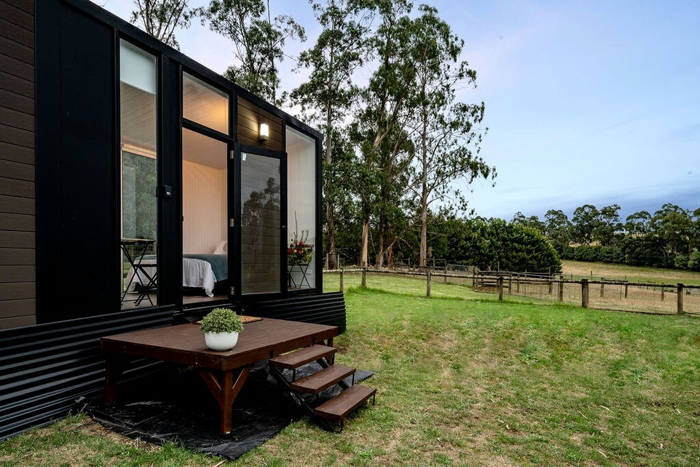 Una casa moderna con un patio que incluye una mesa y bancos de madera, rodeado por un jardín cercado con árboles.