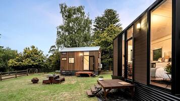 A modern cabin with a wooden exterior, a patio area with a table and benches, and a large window.