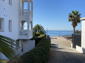 Eine Küstenstraße mit einem weißen Gebäude links, einer Palme und freiem Blick auf das Meer.