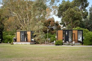 Two modern, compact cabins with large windows, situated in a grassy area with trees in the background.