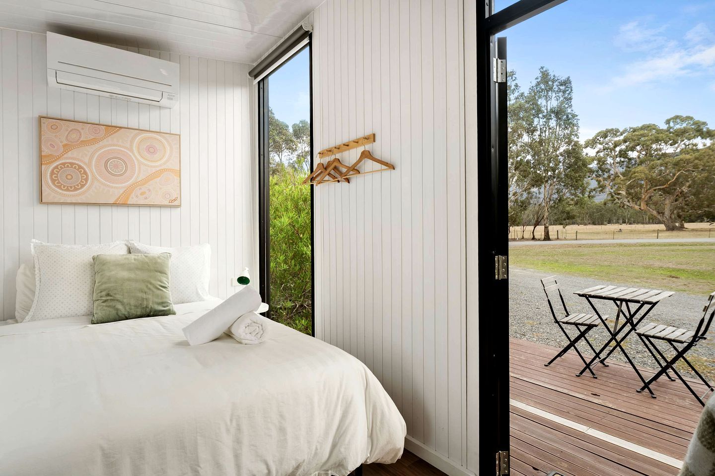 A bedroom with a bed, a wall-mounted air conditioner, a wooden hanger, and a view of an outdoor area with a table and chairs.