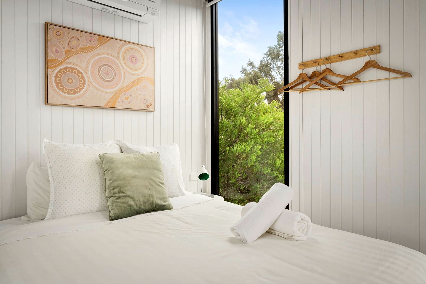 A bedroom with a bed, pillows, a painting, a window, and a wooden hanger rack.