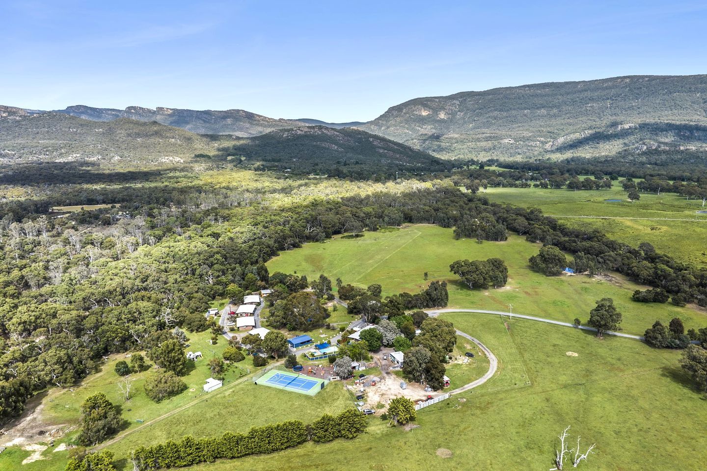 Vista aérea de una propiedad rural con cancha de tenis, rodeada de campos verdes y árboles, con montañas al fondo.