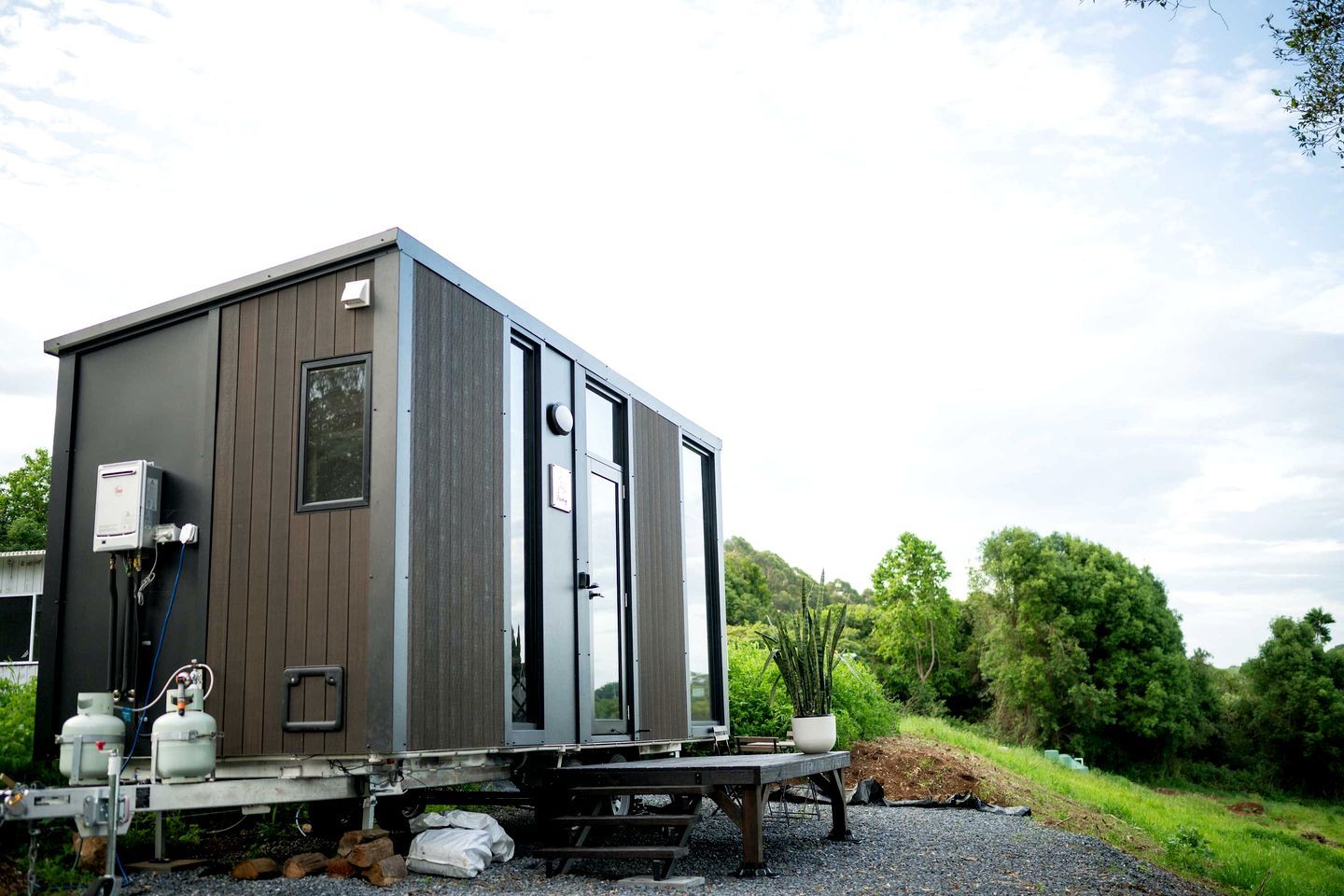 A compact, modern mobile home with a flat roof, a small window, and a door. It is parked on a gravel surface with a green landscape in the background.