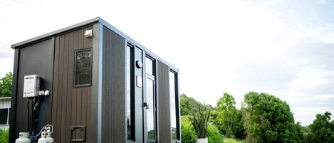 A compact, modern mobile home with a flat roof, a small window, and a door. It is parked on a gravel surface with a green landscape in the background.