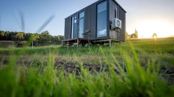 Una estructura moderna de un solo piso con un gran ventanal, un equipo de aire acondicionado y un techo metálico, ubicada en un campo cubierto de hierba.