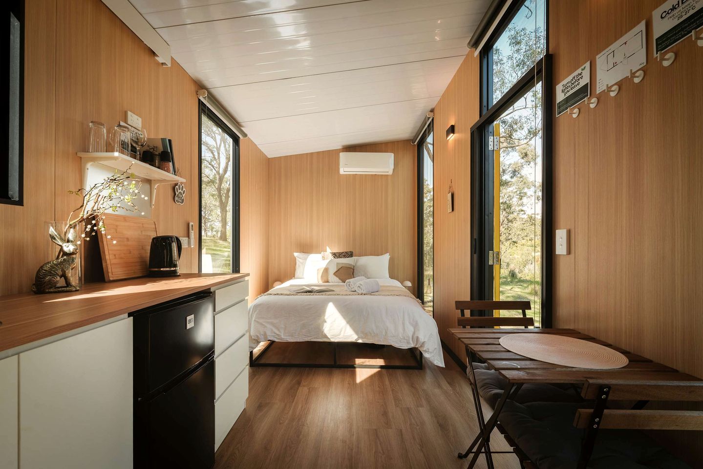 Un dormitorio moderno con techo de madera, cama con ropa de cama blanca, escritorio con silla y ventanas amplias que ofrecen vistas al exterior.