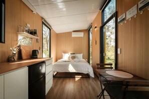 Un dormitorio moderno con techo de madera, cama con ropa de cama blanca, escritorio con silla y ventanas amplias que ofrecen vistas al exterior.