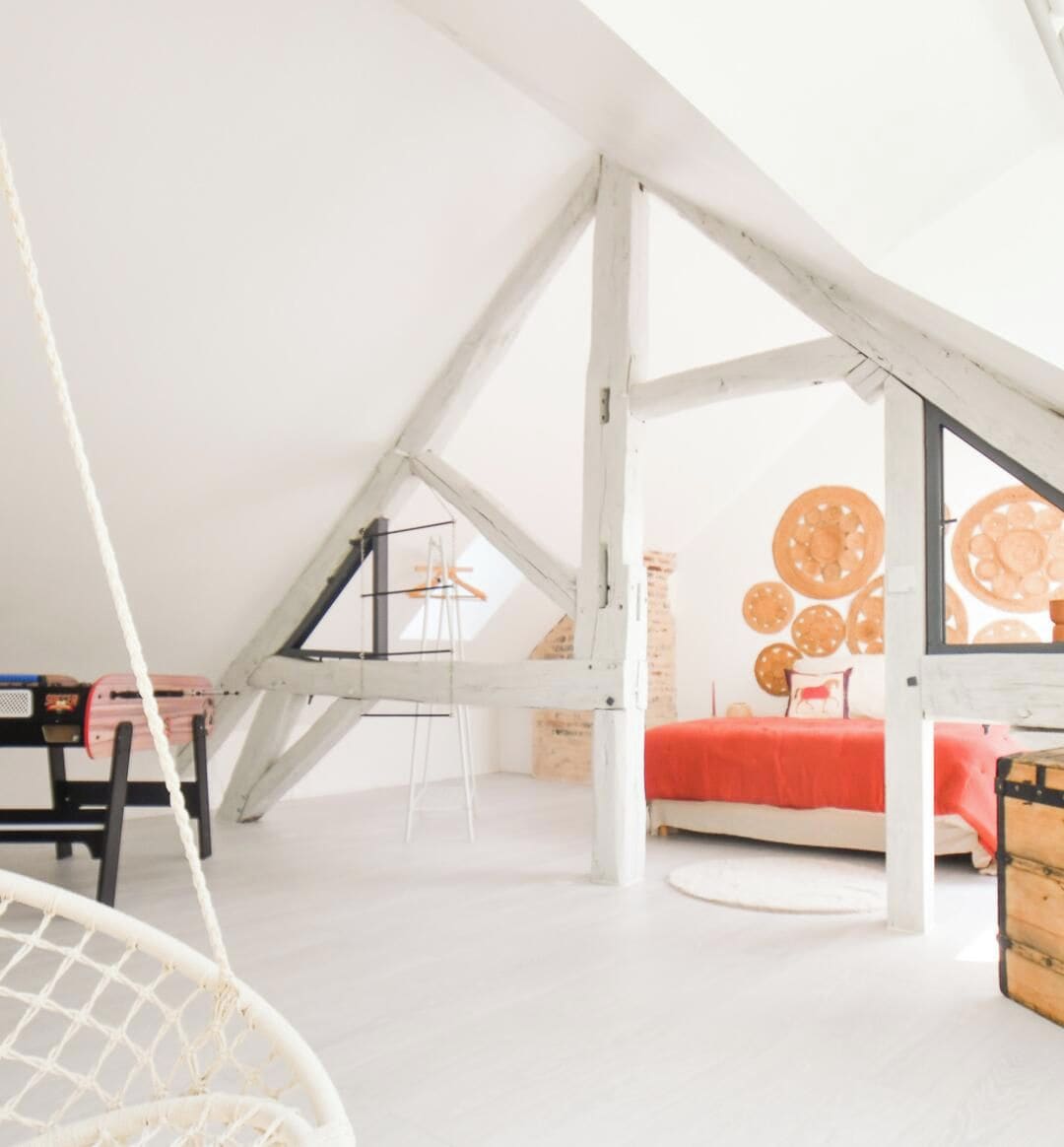 Een slaapkamer in een loft met een schuine kap, een bed met een rode sprei en een hangstoel.