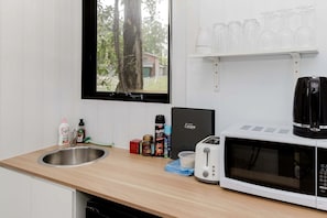 Cozinha com pia de aço inoxidável, micro-ondas, torradeira e prateleira com copos.