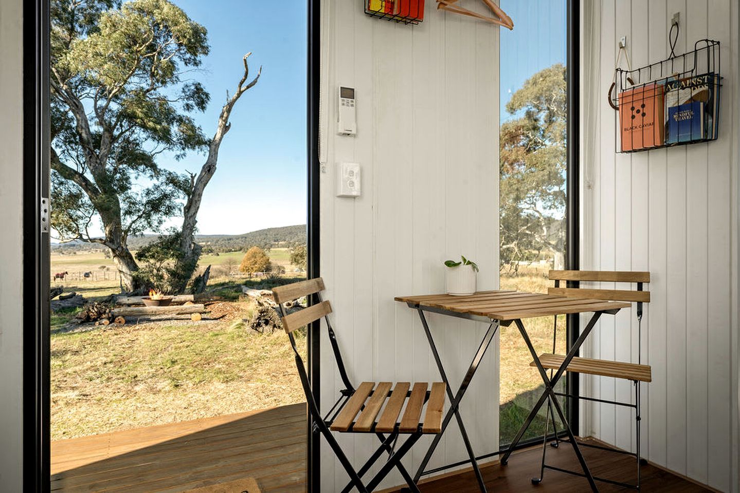 A room with a wooden table and chairs, a small potted plant, and a view of a rural landscape with trees and a fence.