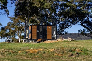 Una cabaña moderna y compacta con un ventanal grande y una puerta, ubicada en un campo cubierto de césped con árboles al fondo.
