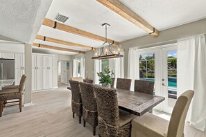 Ein großzügiger Essbereich mit einem großen Holztisch und gewebten Stühlen, einer Decke mit freiliegenden Balken und Blick auf einen Swimmingpool durch die Glasdoor.