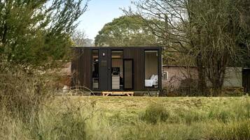 Une petite cabane de couleur sombre, avec une chambre visible et un petit coin cuisine, située près d’une étendue d’eau, avec des arbres en arrière-plan.