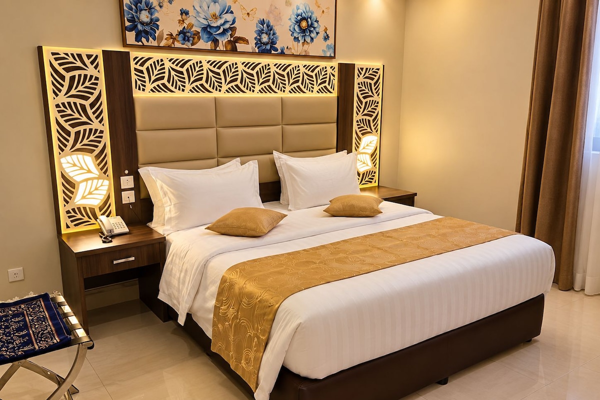 A hotel room with a large bed, a bedside table with a phone, a decorative headboard, and a painting on the wall.