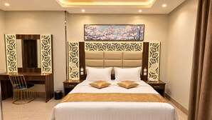 Quarto de hotel com uma cama grande, uma escrivaninha e uma cadeira. Há um quadro na parede e um painel decorativo acima da cama.