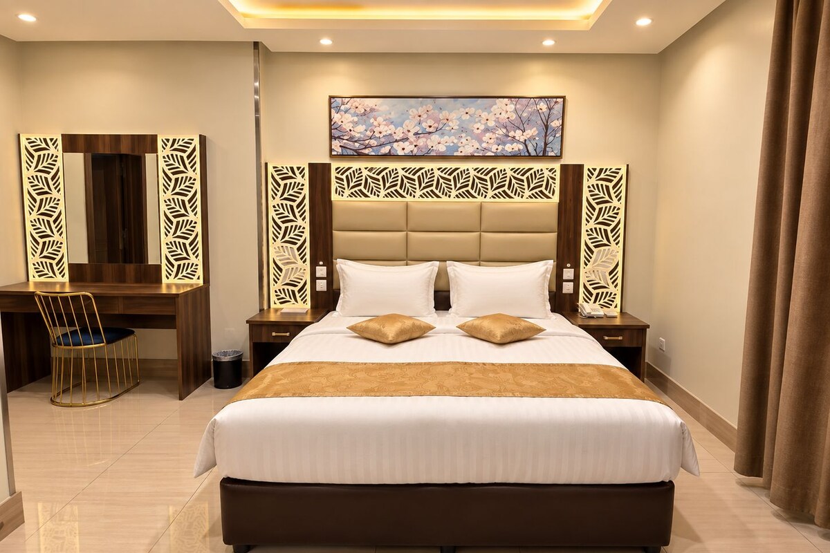 A hotel room with a large bed, a desk, and a chair. There is a painting on the wall and a decorative panel above the bed.