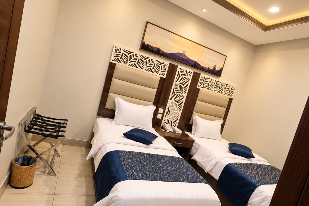 A hotel room with two beds, a wooden headboard, a wall-mounted phone, a small trash bin, and a picture of mountains.