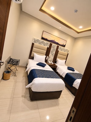 A hotel room with two beds, a wooden headboard, a wall-mounted phone, a small trash bin, and a picture of mountains. - Manazelna serviced apartments- economic (Jeddah)