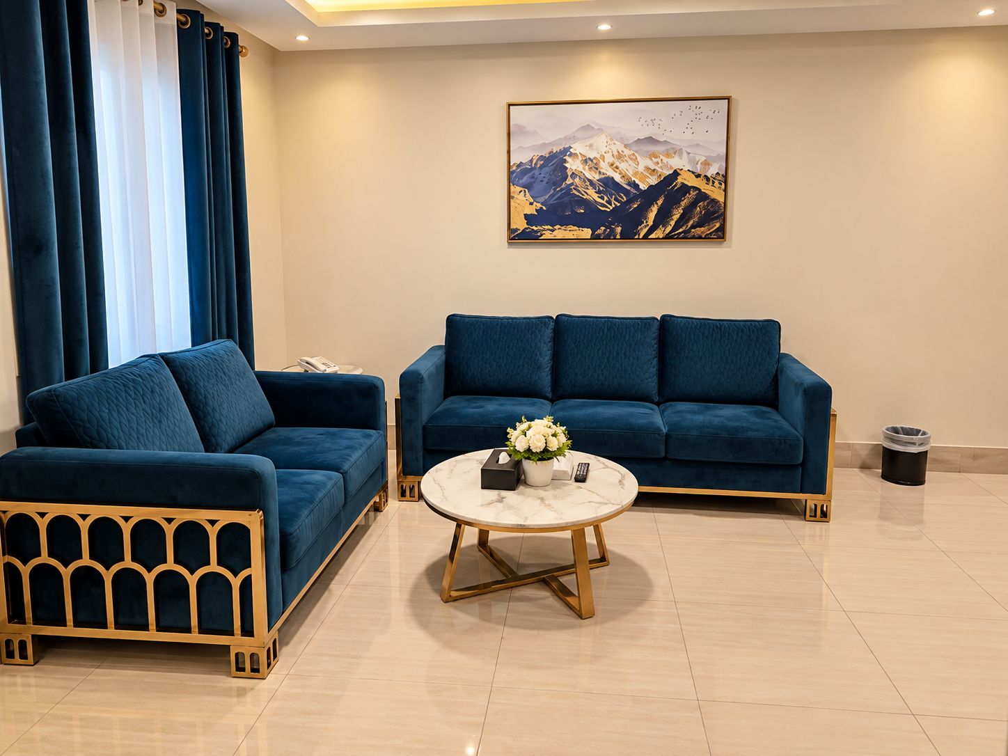 A modern living room with a blue sofa set, a coffee table, and a painting of mountains on the wall.
