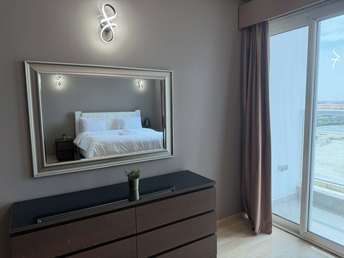 A bedroom with a large mirror, a bedside table with a plant, and a view through a large window.