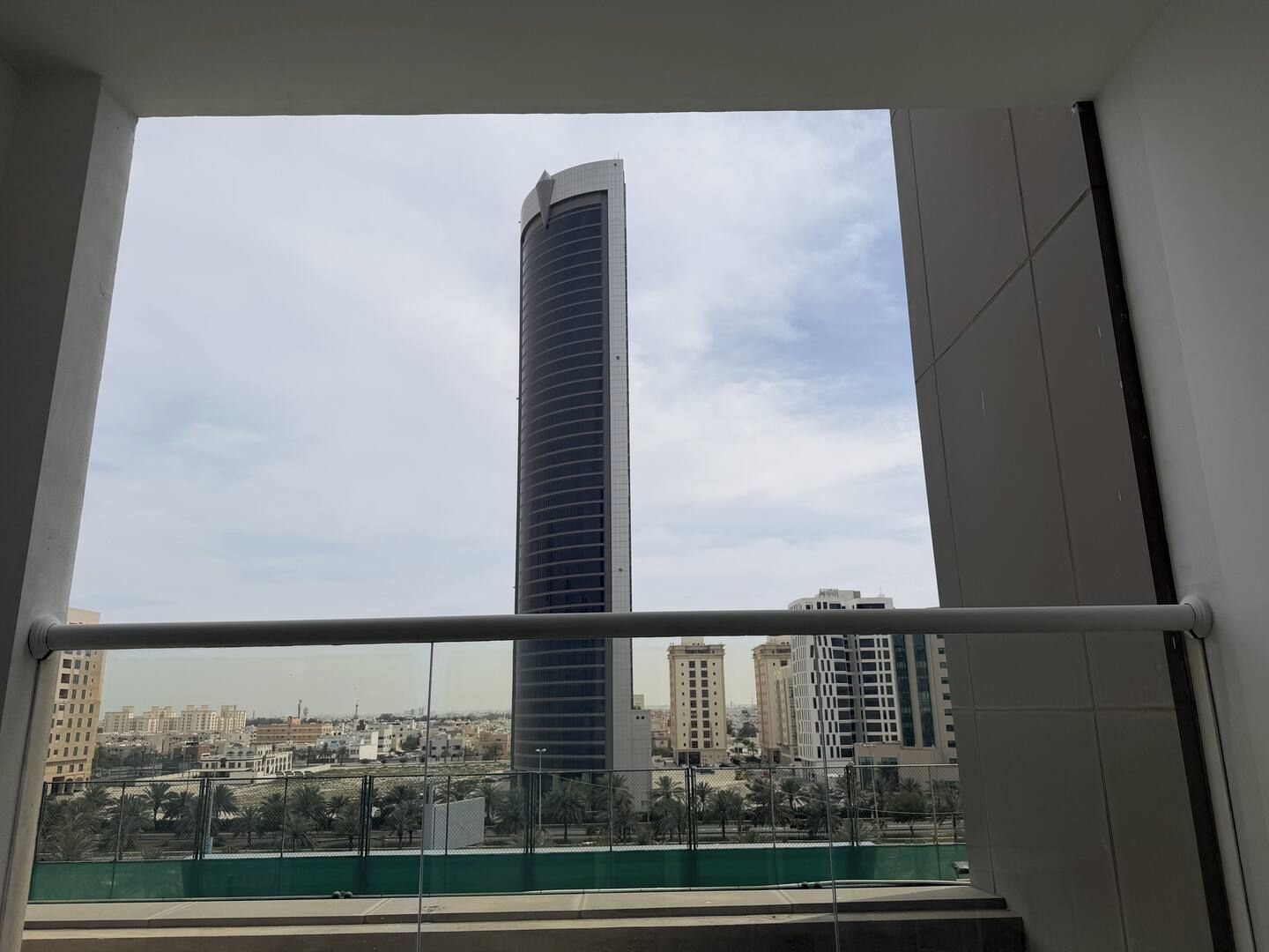 A view from a window with a metal railing, overlooking a tall building and a cityscape with multiple buildings and palm trees.