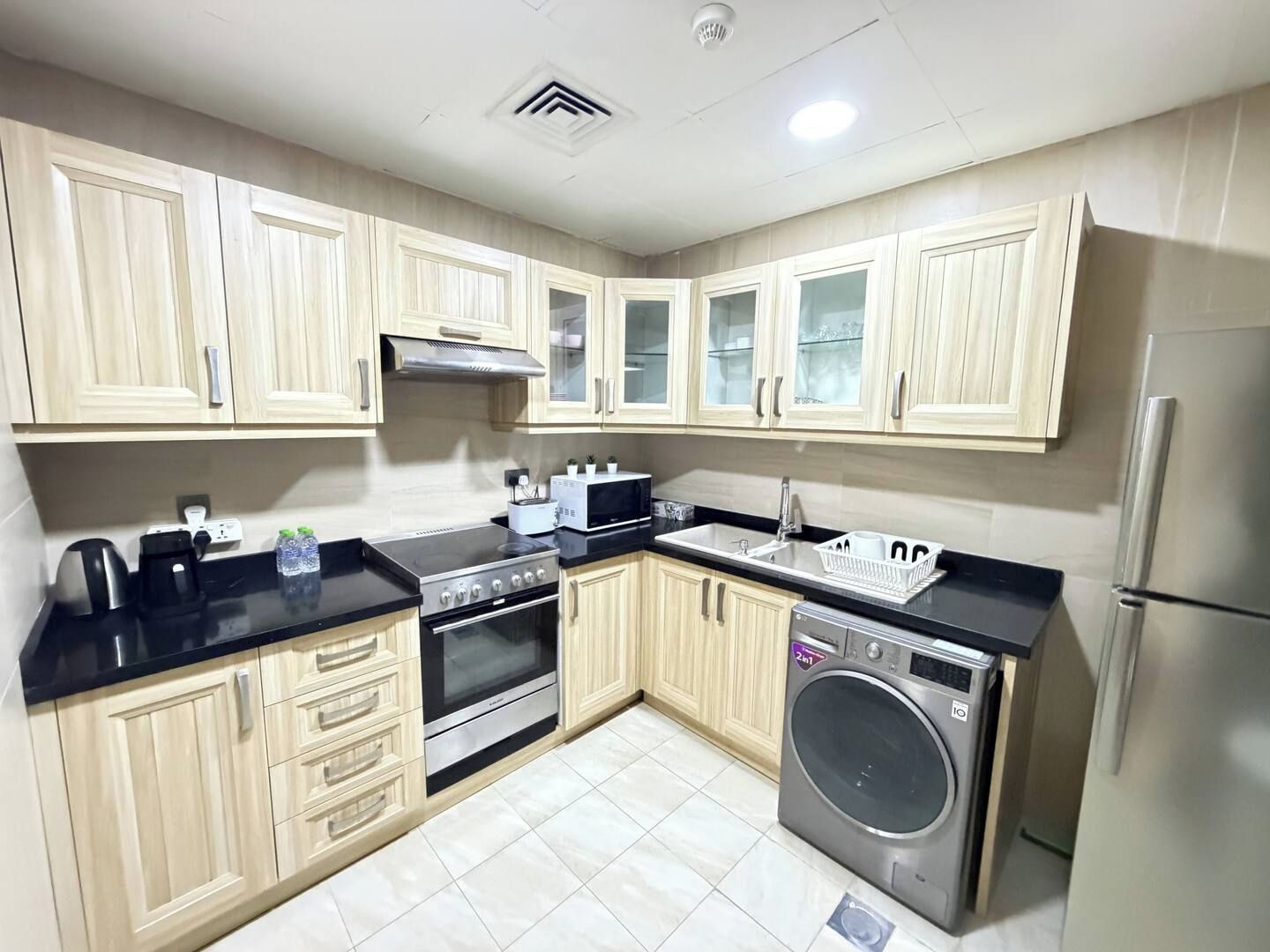 A compact kitchen with light wood cabinets, a stainless steel stove, a microwave, a sink, and a built-in washing machine.