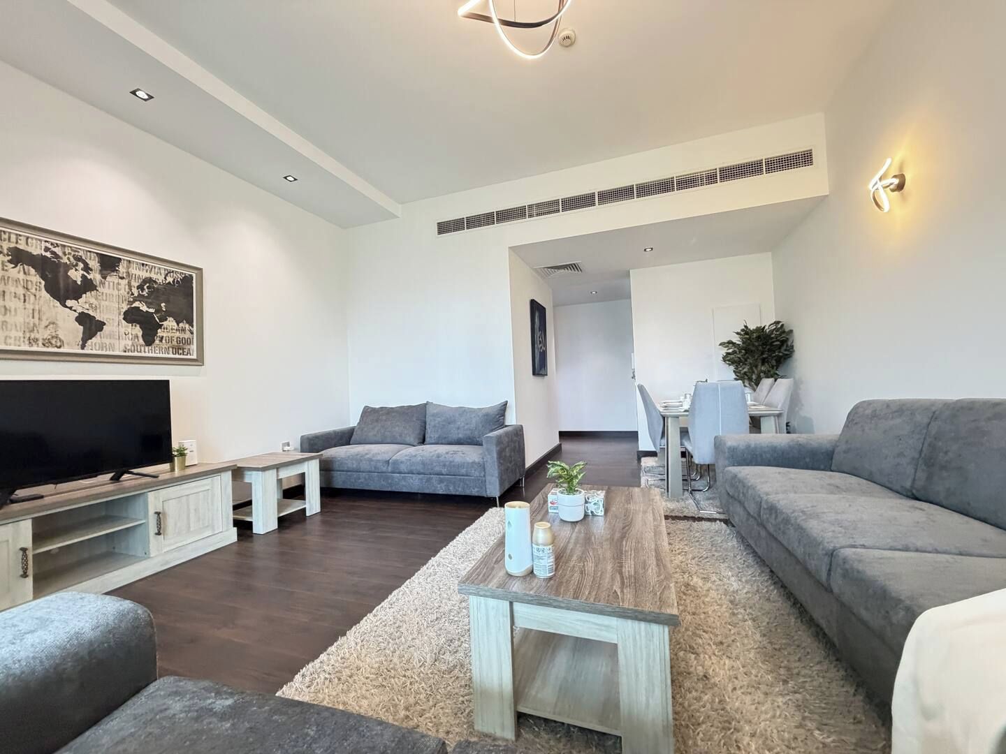 A modern living room with a flat-screen TV, grey sofas, a wooden coffee table, and a dining area with chairs and a table.