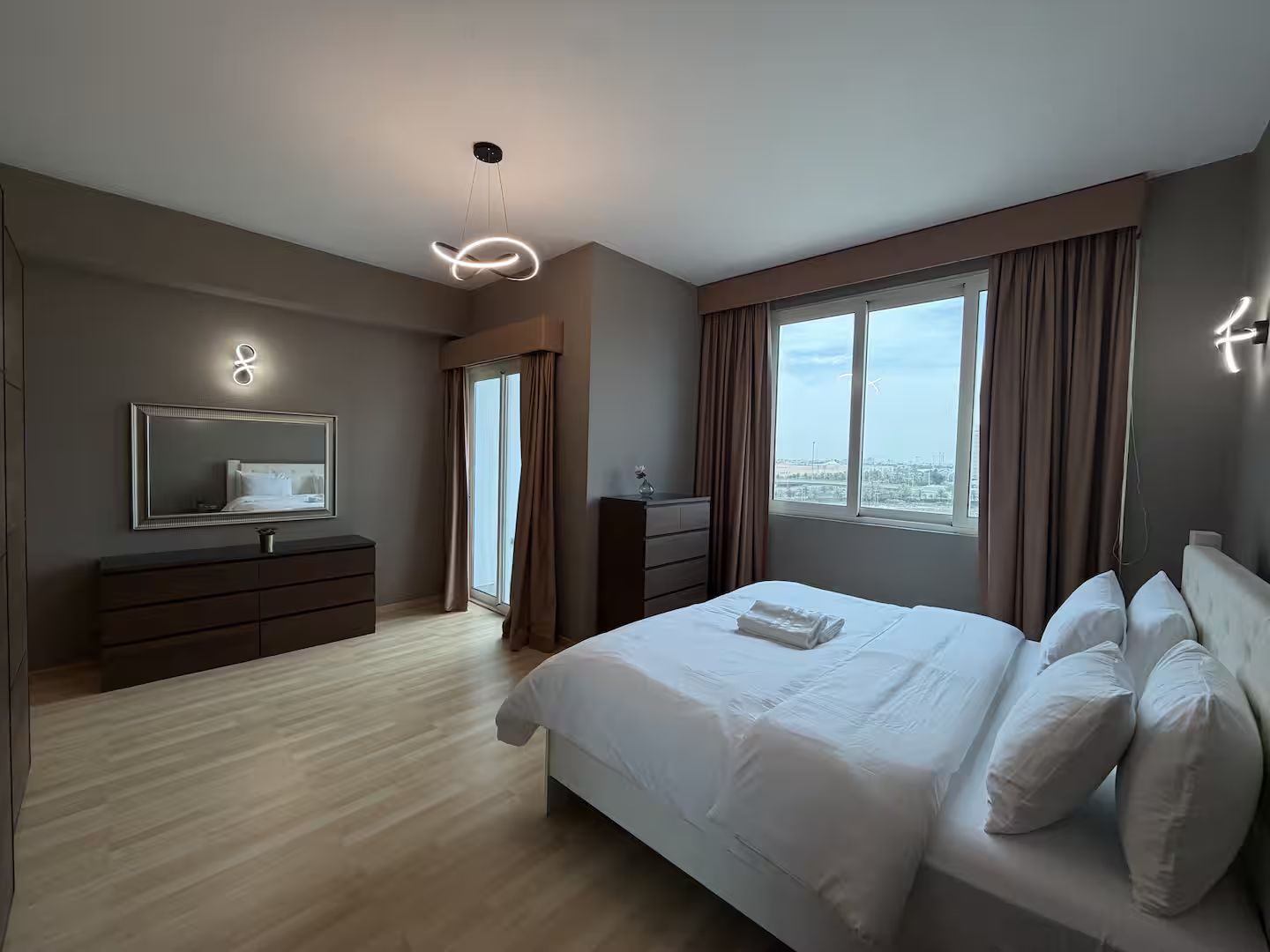 A modern hotel room with a large bed, a dresser, a mirror, and a view of the city through the window.