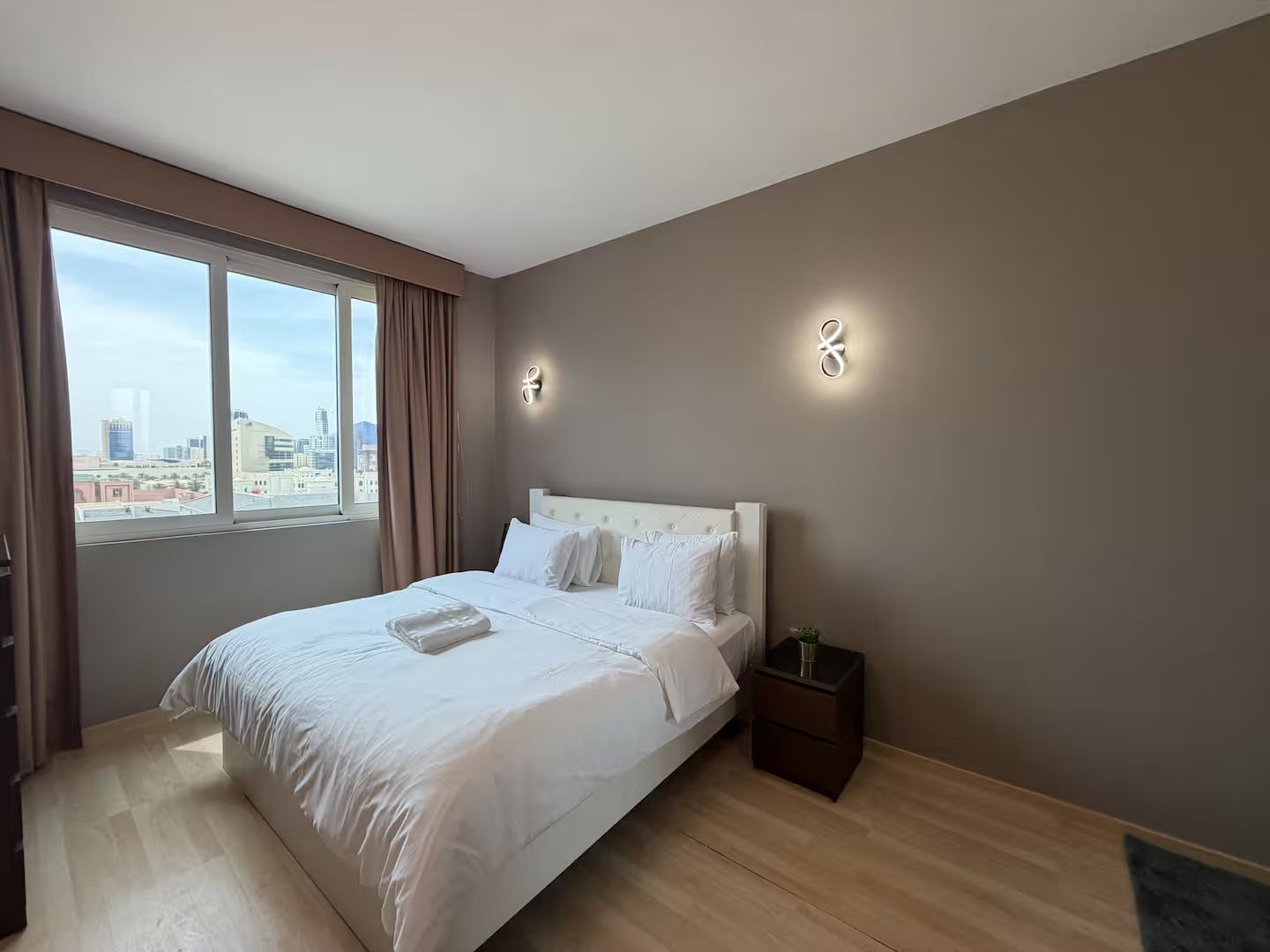 A hotel room with a large bed, a nightstand, a window with curtains, and a city view.