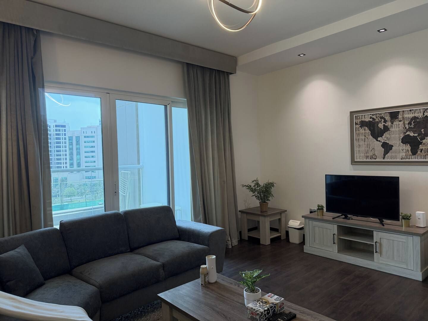 Een moderne woonkamer met een grijze bank, een flatscreen televisie op een houten voetstuk, een wereldkaart aan de muur, en een balkon met gordijnen.