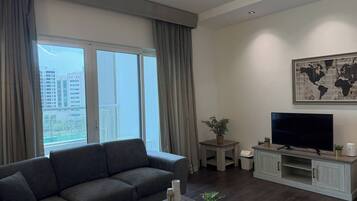 Een moderne woonkamer met een grijze bank, een flatscreen televisie op een houten voetstuk, een wereldkaart aan de muur, en een balkon met gordijnen.