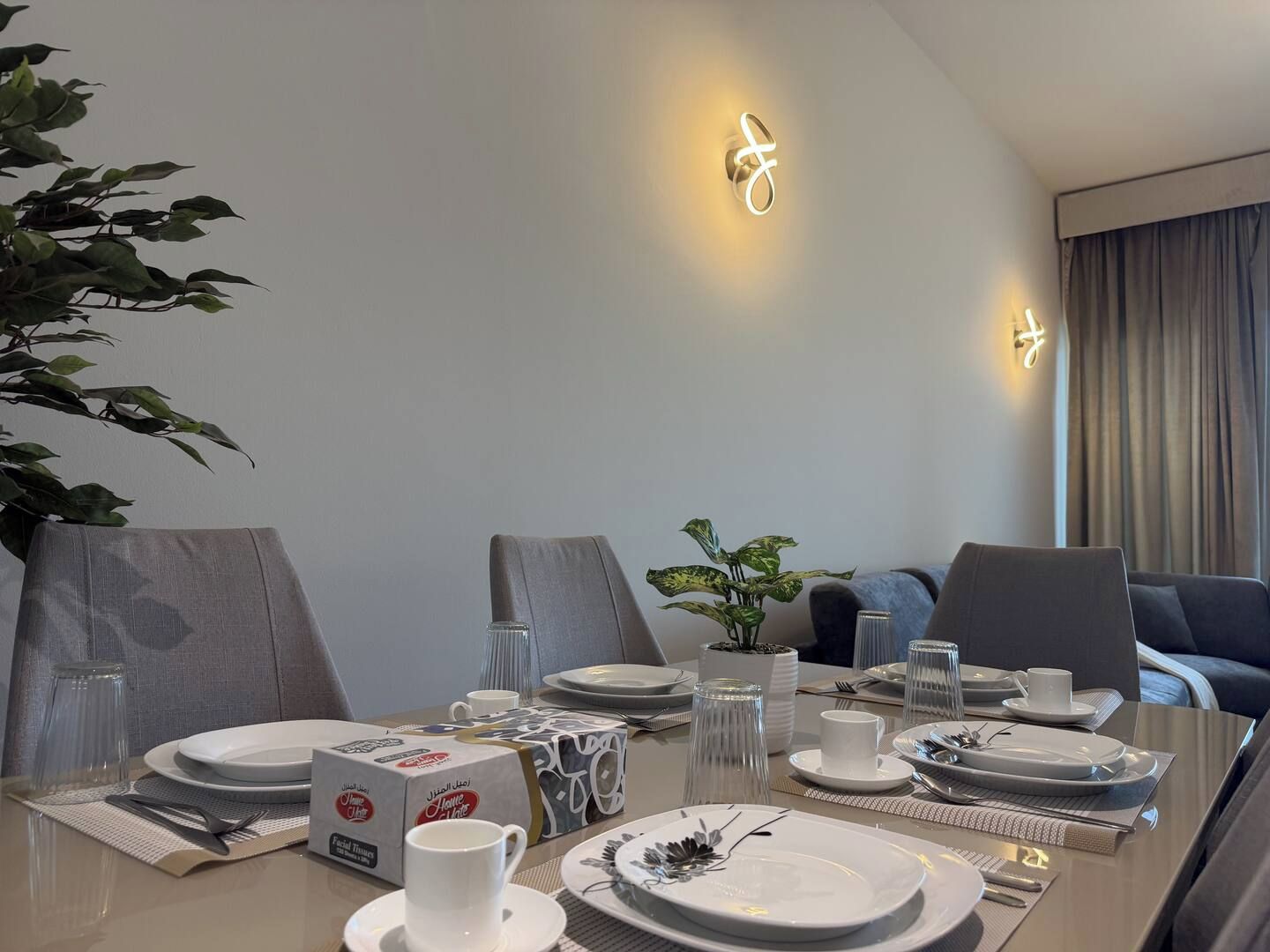 A modern dining area with a table set for four, including plates, cutlery, and a vase with flowers.