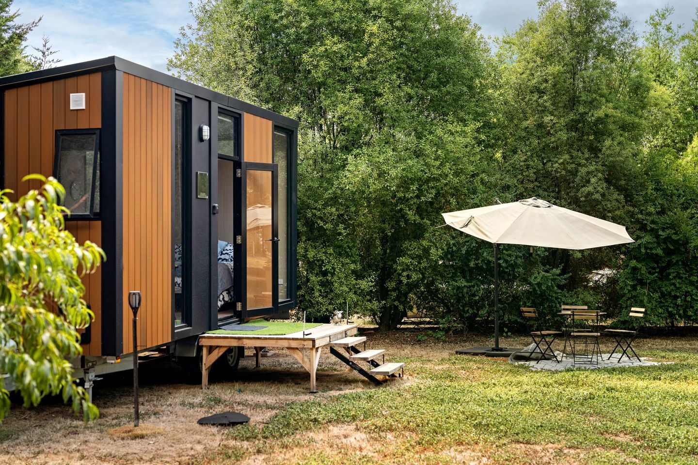 A modern, compact cabin with a wooden exterior and a sliding door, situated in a lush garden with a picnic table and umbrella.
