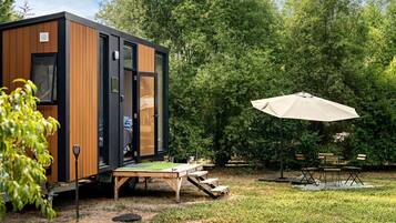 A modern, compact cabin with a wooden exterior and a sliding door, situated in a lush garden with a picnic table and umbrella.
