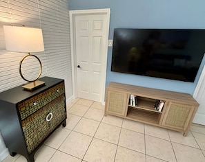 A room with a television mounted on the wall, a small cabinet with a glass top, and a door with a white frame. - Pompano Vibes Beach Apartments - Unit 1 "Ocean Breeze" (Pompano Beach)