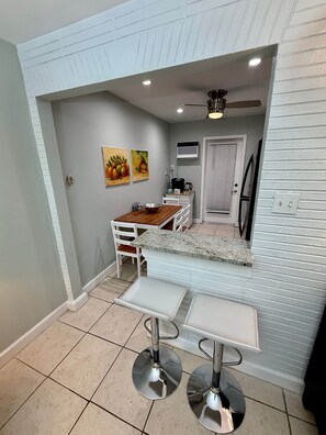 A compact kitchen with a dining area, featuring a white fridge, a coffee maker, and a ceiling fan. - Pompano Vibes Beach Apartments - Unit 1 "Ocean Breeze" (Pompano Beach)