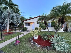 A residential property with a well-maintained lawn, various plants, and a house with a red roof. - Pompano Vibes Beach Apartments - Unit 1 "Ocean Breeze" (Pompano Beach)