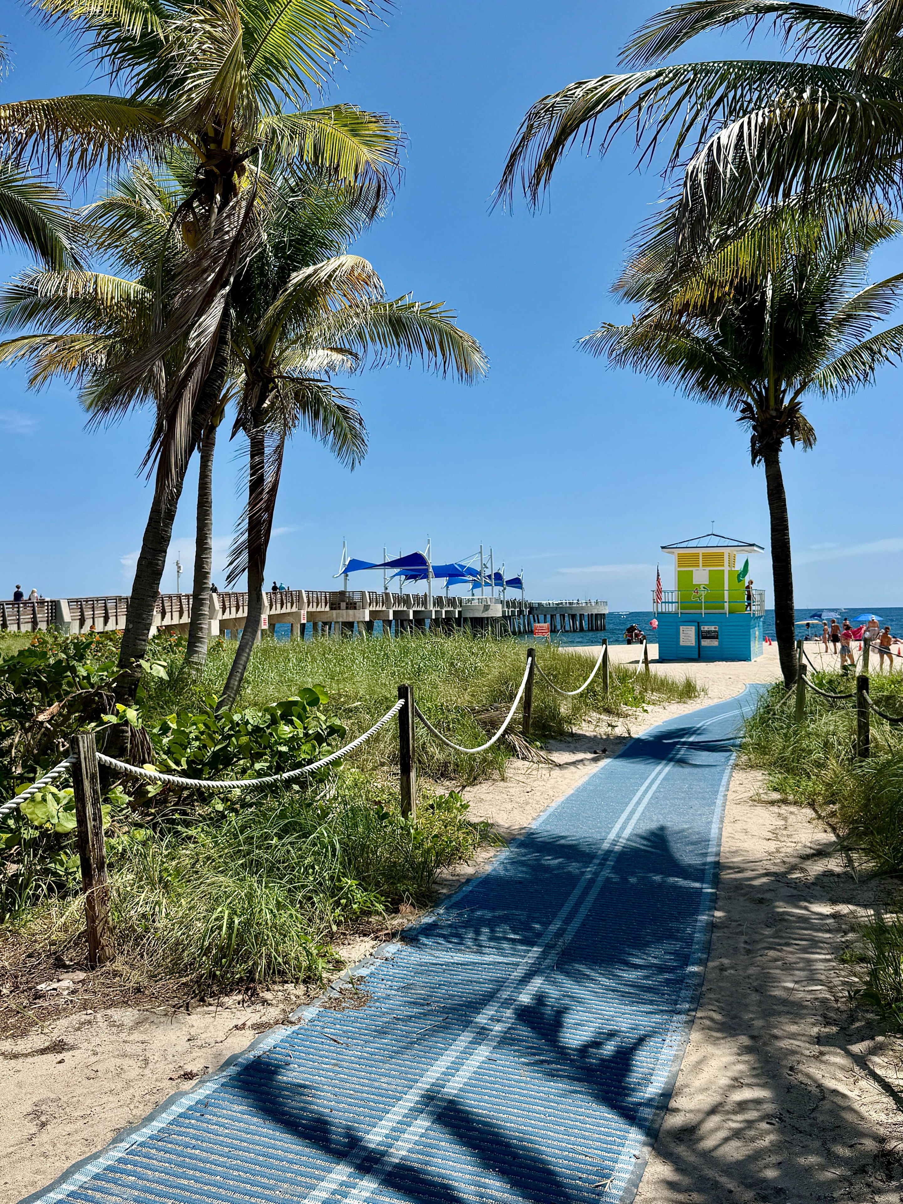 A beach path with palm trees, a lifeguard tower, and a pier with umbrellas.