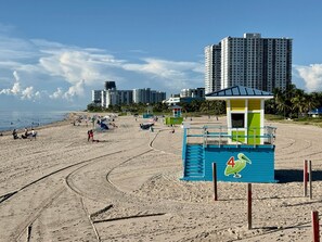 A beach with a lifeguard tower, sandy beach, and high-rise buildings in the background. - Pompano Vibes Beach Apartments - Unit 2 "Coral Reef" (Pompano Beach)