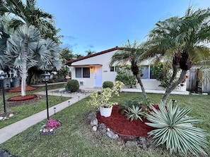 A residential property with a well-maintained lawn, various plants, and a house with a red roof. - Pompano Vibes Beach Apartments - Unit 2 "Coral Reef" (Pompano Beach)