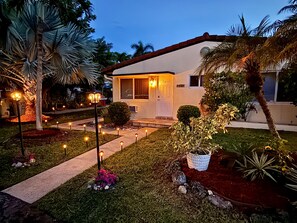 A two-story house with a well-maintained lawn, a pathway leading to the front door, and a small garden with various plants. - Pompano Vibes Beach Apartments - Unit 3 "Pelican's Nest" (Pompano Beach)