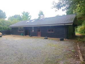 Une cabane en rondins à deux étages, avec un toit en tuiles, un porche à l’avant et une allée gravillonnée.