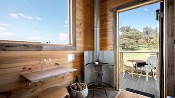 Un interior de madera con chimenea, un banco de madera, una mesa con vasos y vistas al exterior.