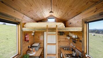 A compact cabin with a loft bed, kitchenette, and a view of a grassy field.